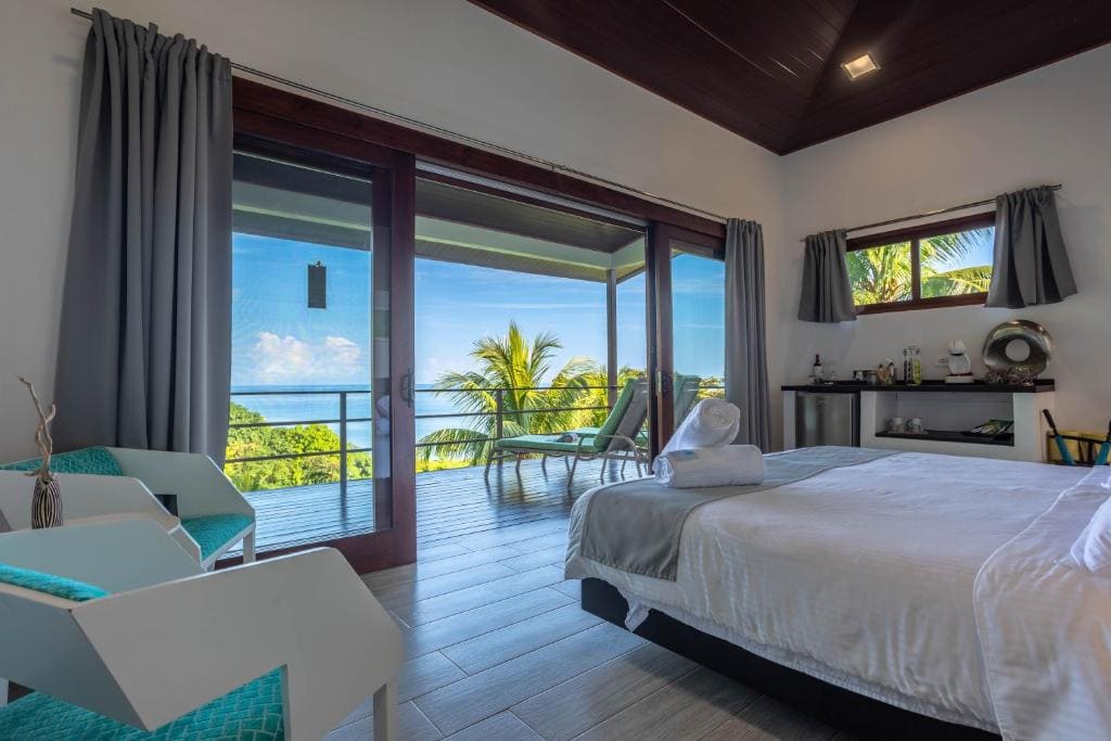 Modern hotel room with a large bed, teal and white chairs, and glass doors opening to a balcony with lounge chairs, overlooking palm trees and an ocean view under a blue sky.