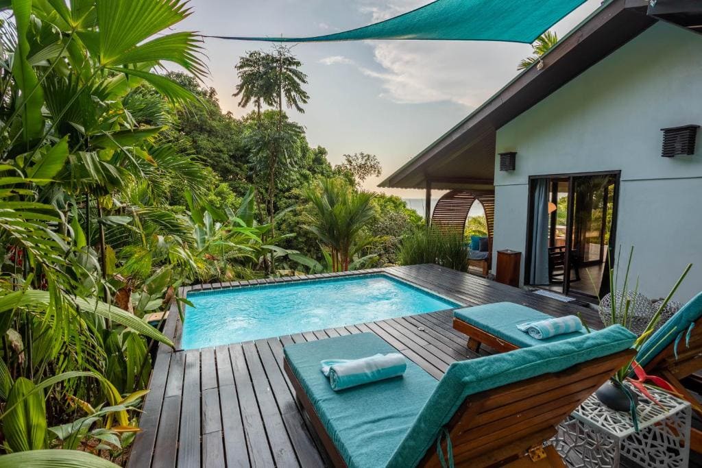 Luxury tropical villa with a private pool, wooden deck, and two lounge chairs with teal towels, surrounded by lush green plants and trees, with a partial ocean view in the background.
