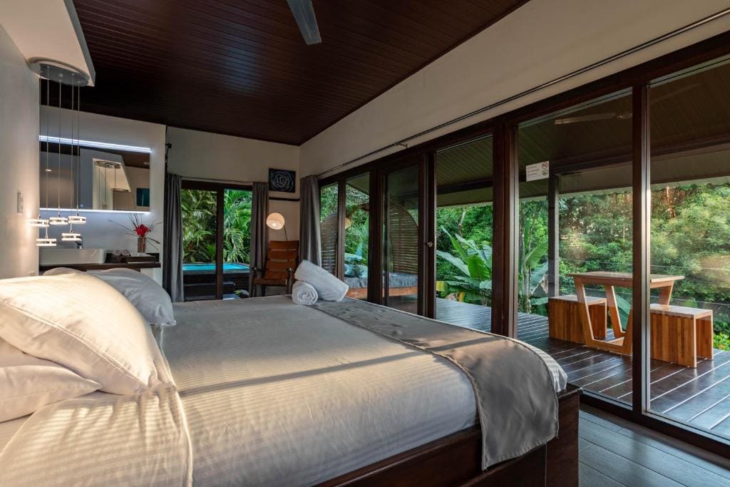 A modern bedroom with a large bed, white bedding, and glass doors opening to a wooden deck with a table and chairs, surrounded by lush green trees and plants. Natural light fills the room.