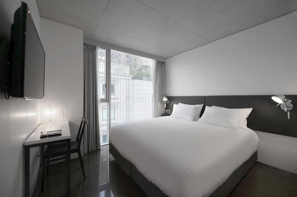 Minimalist hotel room with a large double bed, white bedding, wall-mounted reading lights, a desk and chair, a flat-screen TV, and a window with sheer curtains letting in natural light.