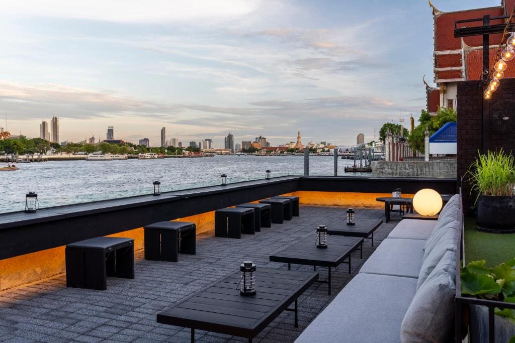 A rooftop terrace beside a river with black tables and benches, modern lanterns, and soft lighting. The city skyline and water are visible in the background under a partly cloudy sky at sunset.