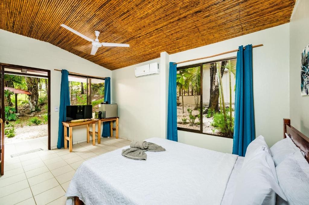 A bright, airy bedroom with a double bed, white bedding, blue curtains, large windows, a wooden ceiling, a ceiling fan, and a small desk with a TV and fridge. Tropical greenery is visible outside.
