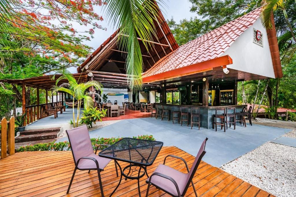 An outdoor restaurant and bar with a red-tiled roof, surrounded by lush greenery. There are tables and chairs on a wooden deck in the foreground and a covered bar area with high stools in the background.