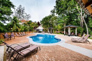 Outdoor swimming pool surrounded by lounge chairs, gazebos, and lush tropical trees, with a house in the background and a partly cloudy sky overhead.