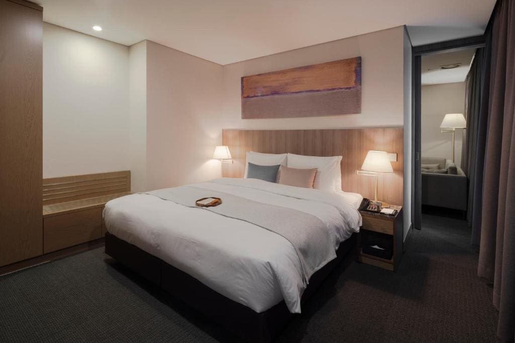 A modern hotel room with a large bed, wooden headboard, side table with a lamp and phone, minimal decor, and neutral-colored walls and bedding; a lamp and chair are visible in an adjacent room through an open doorway.