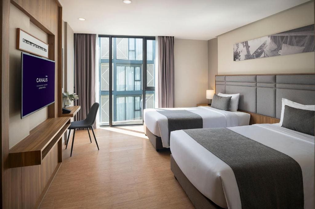 Modern hotel room with two neatly made beds, gray headboards, a desk with a chair, flat-screen TV on the wall, large window with curtains, and light wood floors. The room is bright and tidy.