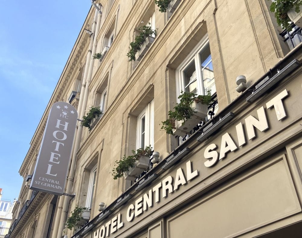 Hotel Central Saint Germain building facade with beige stone exterior, multiple windows with flower boxes, and a vertical sign reading HOTEL Central St Germain in French, seen from a street-level upward angle.
