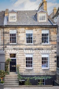 A traditional stone townhouse with three stories and white-trimmed sash windows, featuring a black door with a wreath, greenery by the entrance, and steps leading down to a lower ground floor.