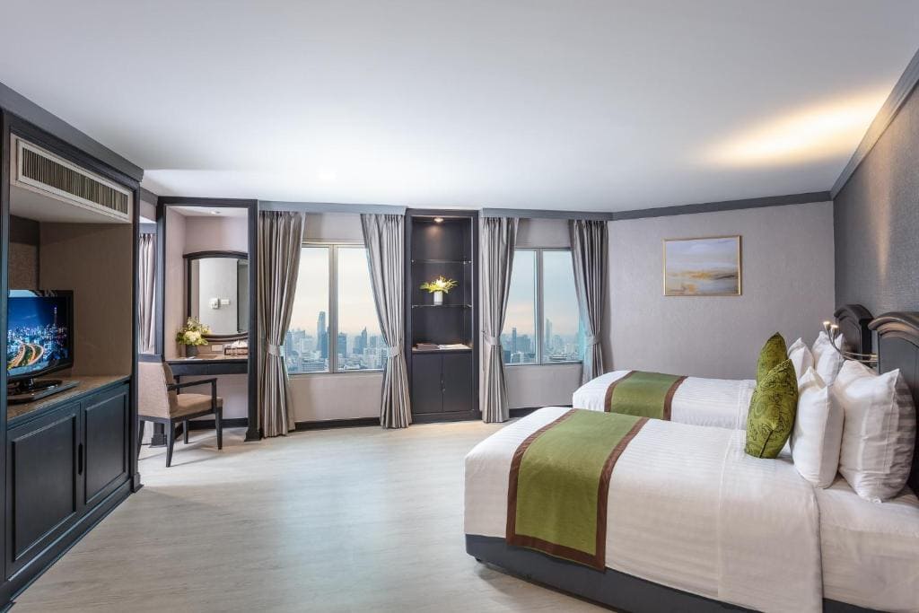 Spacious hotel room with two beds, green accent pillows and runners, a desk by large windows with city views, shelves with decor, a TV, and neutral-colored walls and flooring.