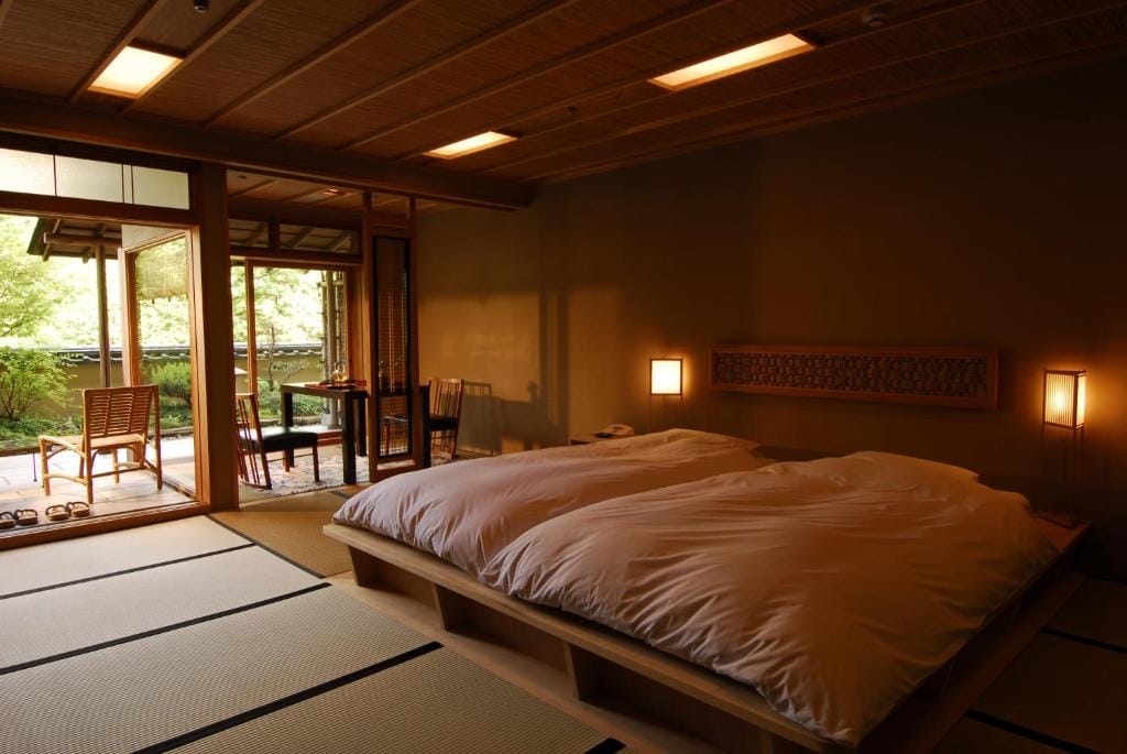 A serene Japanese-style room with two futon beds on a wooden platform, tatami mat flooring, soft lighting, and an open sliding door leading to a garden patio with chairs and lush greenery outside.
