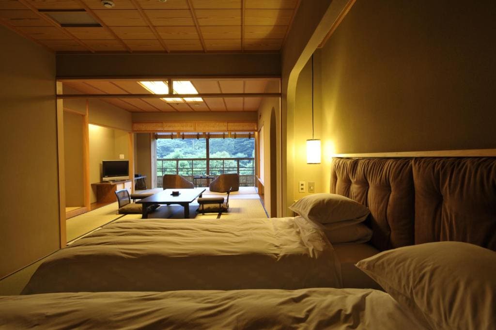 A cozy Japanese-style room with two beds in the foreground, a low table with chairs in the middle, tatami mats, and large windows in the background displaying lush green trees. Warm lighting fills the space.