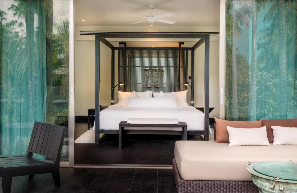 A modern hotel room with a four-poster bed, white bedding, a bench at the foot, turquoise curtains, an outdoor lounge chair, and green trees visible through the glass doors.