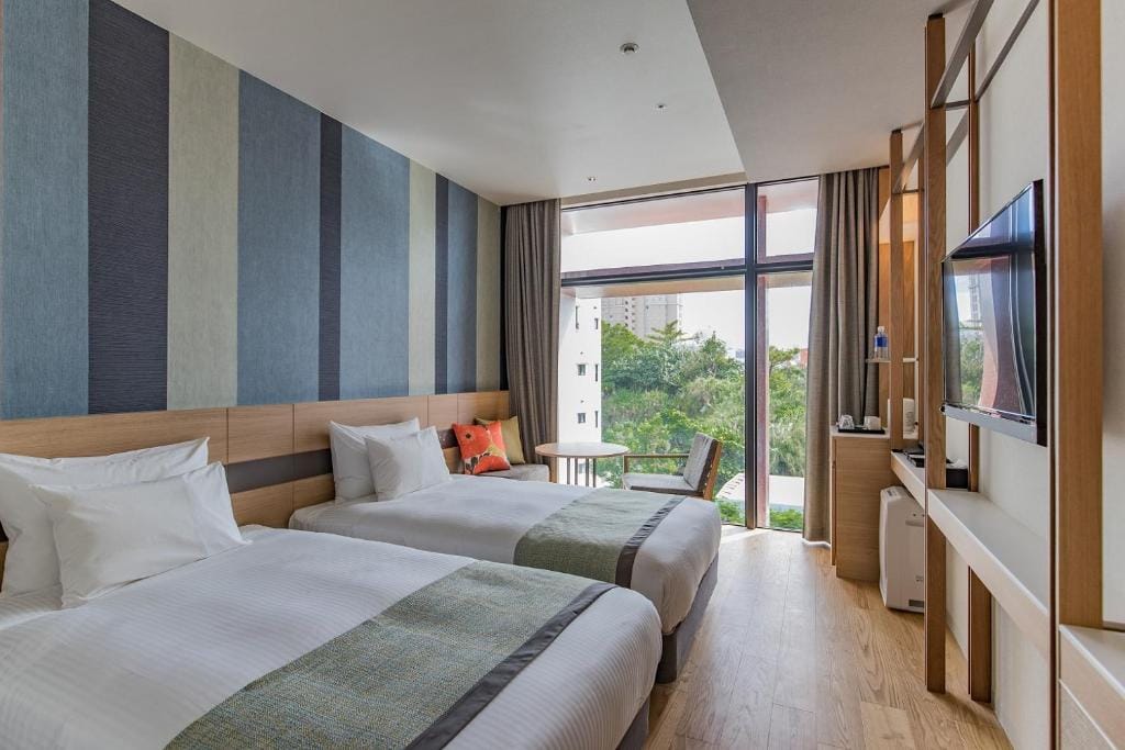 Modern hotel room with two twin beds, striped accent wall, large window with a view of greenery and buildings, a small round table with two chairs, a wall-mounted TV, and light wood furnishings.