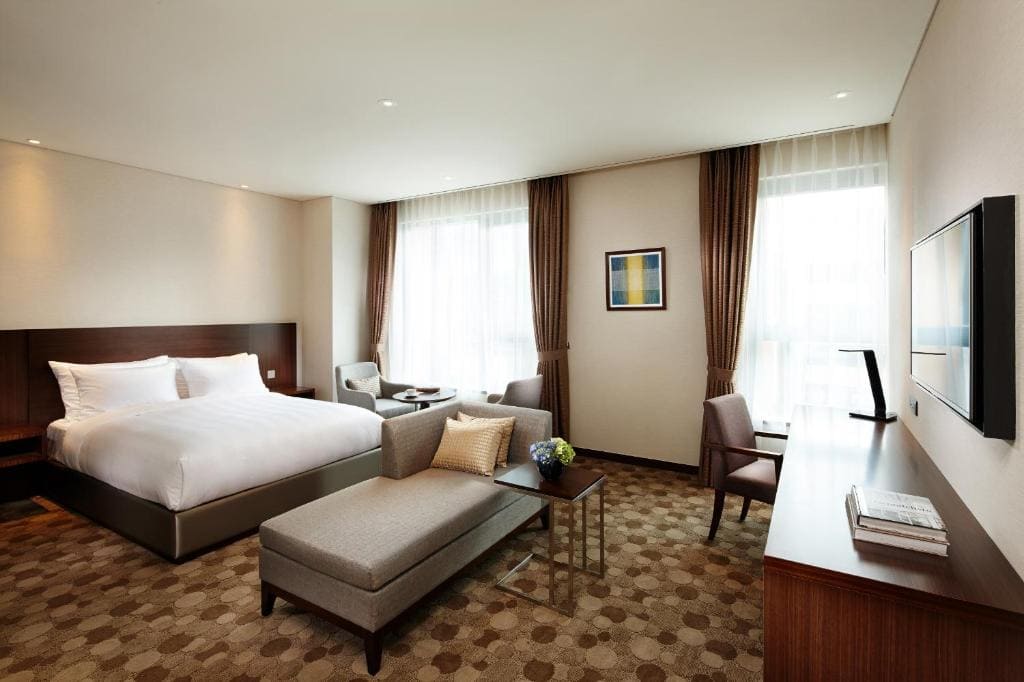 A modern hotel room with a large bed, a gray chaise lounge, a desk with a chair, two armchairs by the window, neutral walls, patterned carpet, and a wall-mounted TV. Natural light fills the room.