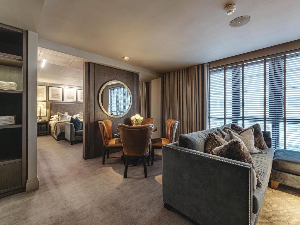 A modern hotel suite with a living area featuring a gray sofa, armchairs around a round dining table, a large circular wall mirror, and a bedroom visible through an open doorway. Floor-to-ceiling windows have horizontal blinds.