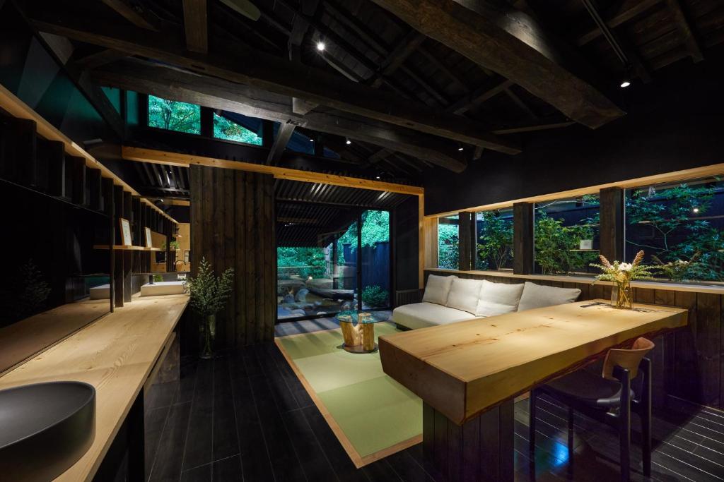 Modern, minimalist living space with wooden beams, open shelving, a light-colored sofa, and large windows overlooking lush greenery; natural light fills the room, highlighting wooden and green elements.
