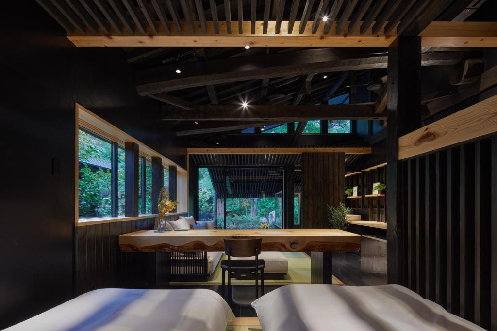 A modern, minimalist bedroom with dark wooden beams, large windows revealing greenery, light wooden accents, a raw-edge wooden desk, a single chair, and shelves along the back wall. Natural light brightens the space.