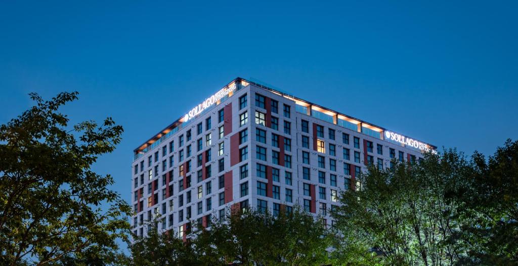 A modern, multi-story building with illuminated signage on the rooftop, seen at dusk against a clear blue sky, with trees in the foreground.