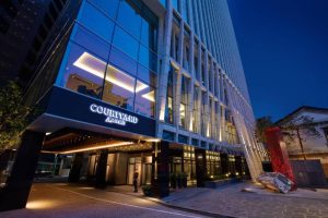 Modern high-rise Courtyard Marriott hotel exterior at dusk, with illuminated sign, glass facade, street lights, and a red sculpture near the entrance. A person stands by the revolving door.