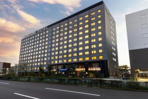 A large, modern Mercure Hotel building with numerous lit windows, standing next to a road at sunset. The sky is partly cloudy and the hotel’s name is displayed prominently at the top.