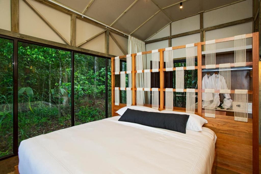A modern bedroom with a large bed, wood and rope headboard, white linens, and a long black pillow. Floor-to-ceiling glass windows reveal lush green forest outside. A shelf with robes and a safe is behind the headboard.