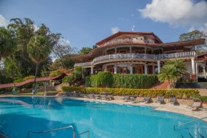 A large, multi-level tropical resort building with balconies overlooks a clear blue swimming pool surrounded by lounge chairs, palm trees, and lush greenery under a sunny sky.