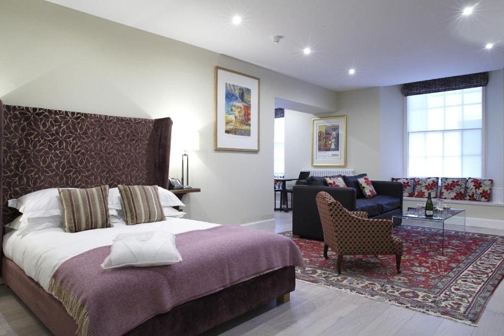 A bright, modern bedroom with a large bed, patterned headboard, and purple bedding. There’s a seating area with a sofa, armchair, coffee table, and colorful rug. Artworks and large windows enhance the room’s airy feel.