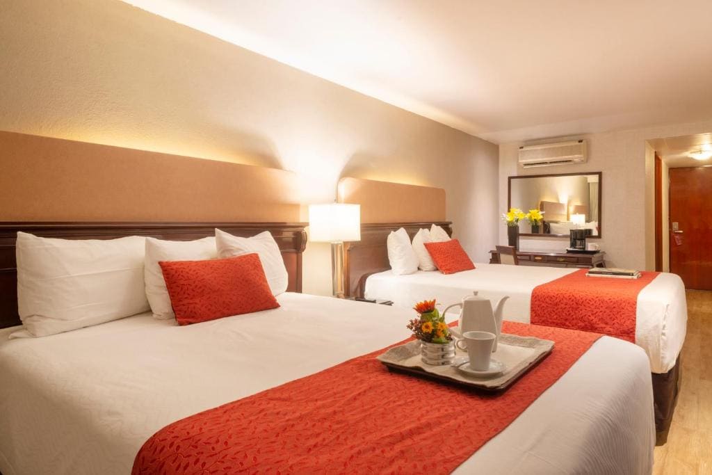 A hotel room with two double beds, each with white linens and red runners and pillows. A tray with a teapot, cups, and flowers sits on one bed. There’s a lamp, mirror, and flowers on a desk in the background.