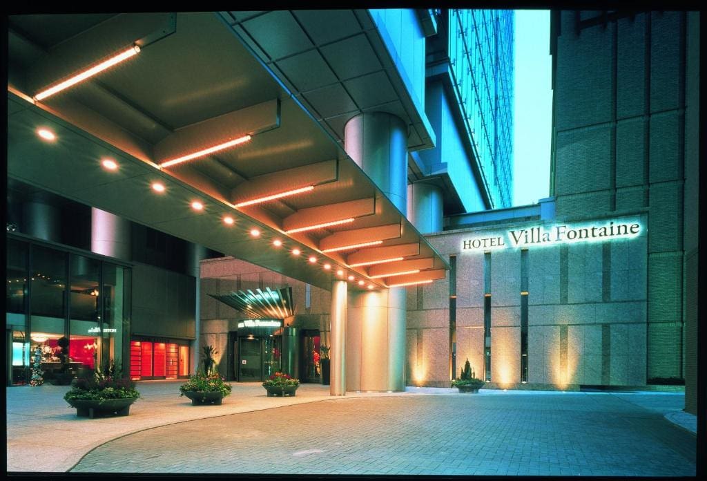 The modern entrance of Hotel Villa Fontaine, featuring a covered driveway lit with warm lights, potted plants, and the hotels name illuminated on the buildings exterior wall.