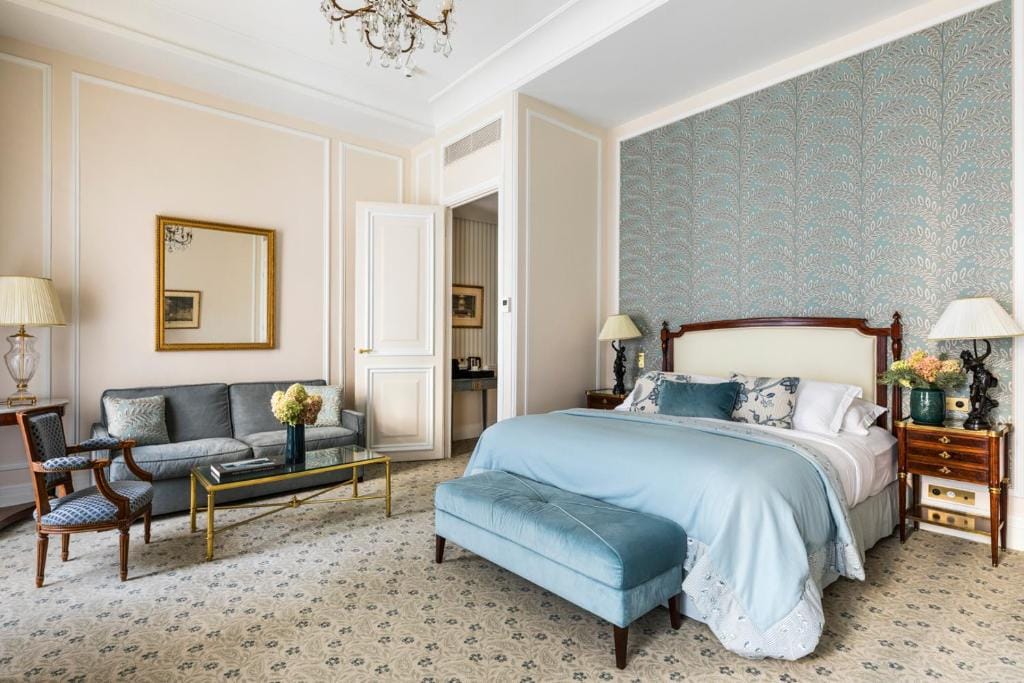 Elegant hotel room with a large bed, blue bench, patterned accent wall, sofa, armchair, coffee table, and two bedside tables with lamps and flowers. A chandelier, wall mirror, and soft carpet complete the decor.