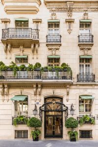 Ornate European building facade with wrought-iron balconies, green plants, and large windows. The entrance features an arched glass door, lantern-style lamps, and symmetrical potted trees on either side.