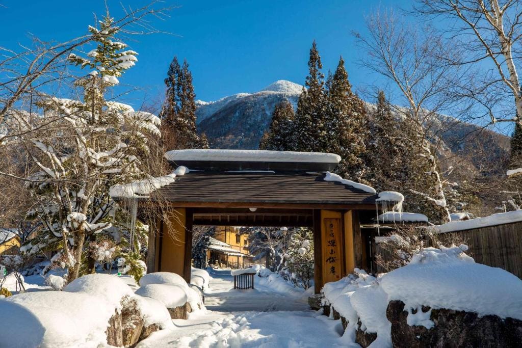 A traditional Japanese gate stands amid snow-covered trees and bushes, with a clear blue sky and a mountain in the background. Sunlight highlights the wintry scene, creating a serene atmosphere.