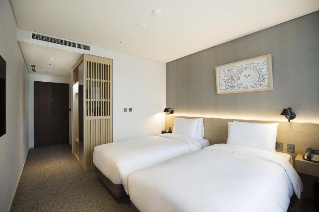 Modern hotel room with two single beds, white bedding, soft lighting, a framed artwork on the wall, and a wooden partition near the entrance; minimalistic and clean design.