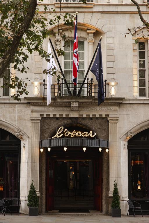 The entrance of a building with Foscar written above the door, three flags above, and two potted plants at the entrance. The building has arched windows and stone detailing.