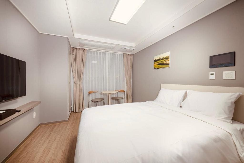 Minimalist hotel room with a large white bed, wall-mounted TV, small table and two chairs by a window with beige curtains, and a landscape picture on a light-colored wall.