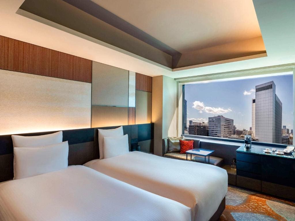 Modern hotel room with two neatly made twin beds, large window showing a cityscape with tall buildings, a cushioned bench, a small table, and contemporary decor in warm tones.
