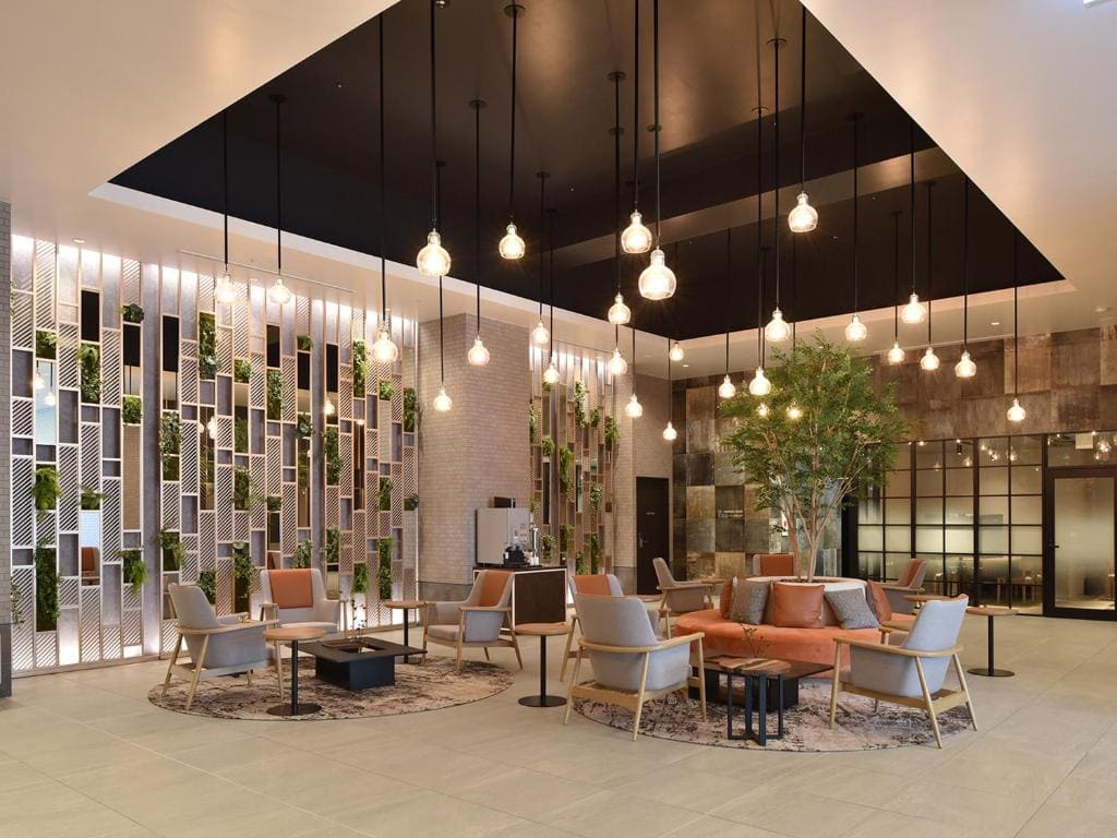 Modern hotel lobby with stylish seating areas, hanging pendant lights, a large central plant, decorative wall with greenery, and neutral-toned furniture arranged on patterned rugs.