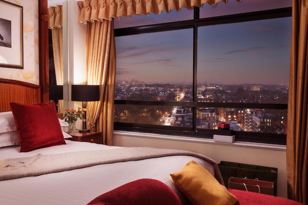 A cozy hotel room with a large bed, red and gold cushions, warm lighting, and a window offering a cityscape view at dusk with twinkling lights under a colorful sky.