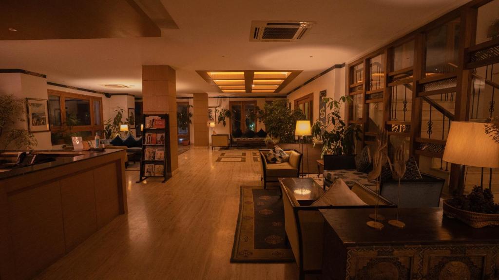 A warmly lit hotel lobby with sofas, potted plants, bookshelves, and lamps, featuring wooden accents and large windows along the right wall. The reception desk is on the left.