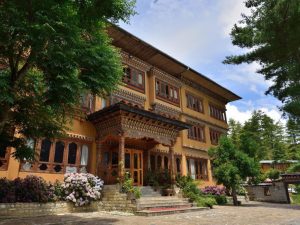 A traditional Bhutanese building with ornate wooden carvings, large windows, and colorful details, surrounded by lush green trees, flowers, and a stone pathway under a partly cloudy sky.