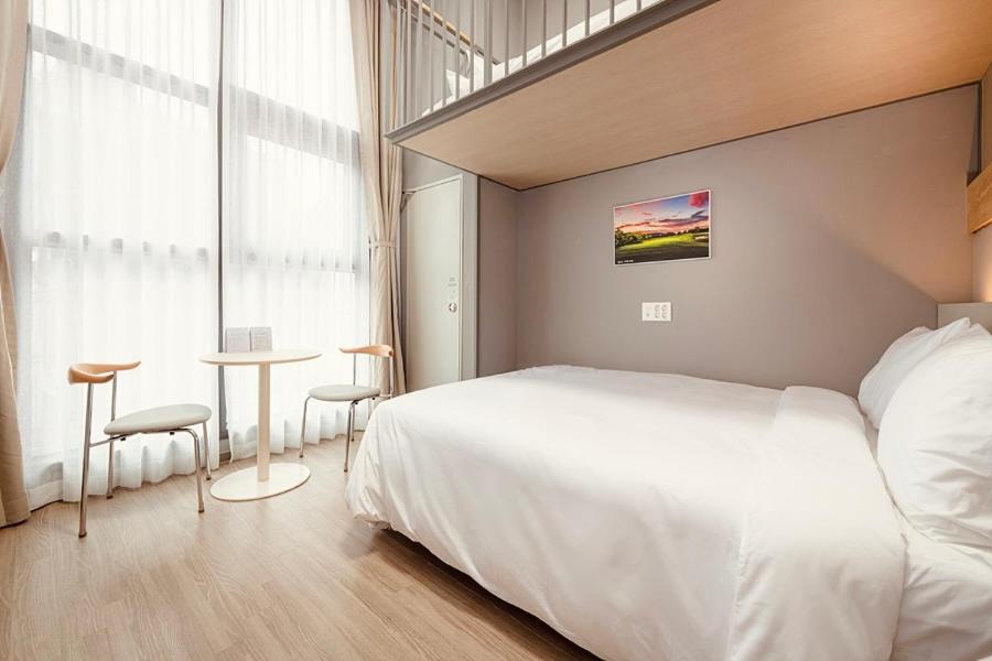A modern, minimalistic hotel room with a large bed, a small round table with two chairs by tall windows with sheer curtains, and a loft space above. The room features light wood floors and neutral-toned walls.
