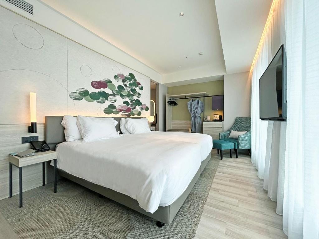 A modern hotel room with two neatly made beds, a wall mural, a robe hanging in an open closet, a turquoise armchair, a large window with sheer curtains, and a flat-screen TV mounted on the wall.