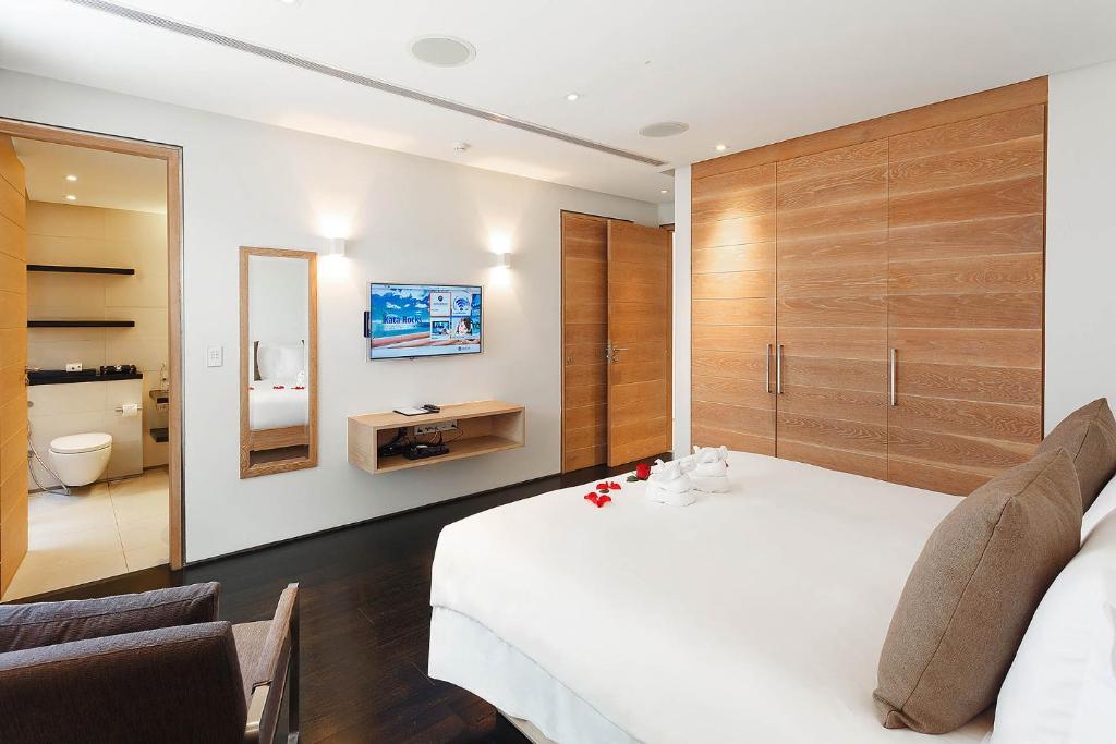 A modern hotel room with a large bed, brown pillows, and towel art on white bedding. There are wooden wardrobes, a wall-mounted TV, a mirror, and an open bathroom with a toilet and shelves visible.