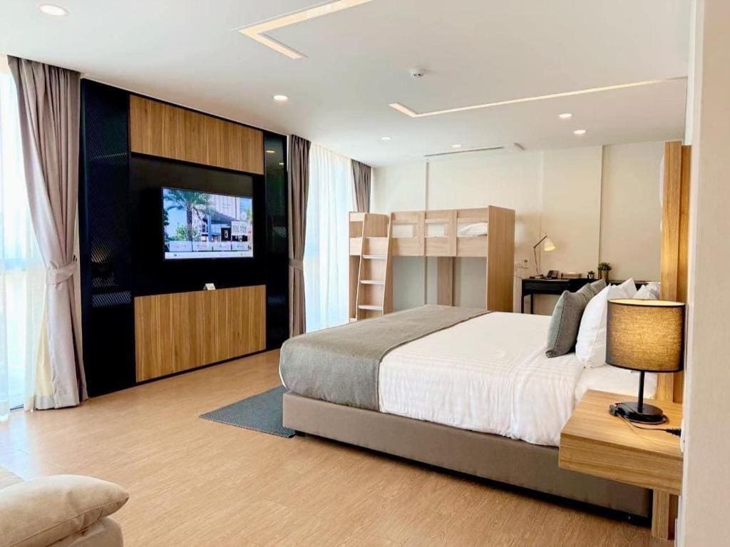 A modern hotel room with a large bed, wall-mounted TV, bunk beds in the corner, a desk with a kettle, and floor-to-ceiling windows with curtains, all decorated in neutral tones and wood accents.