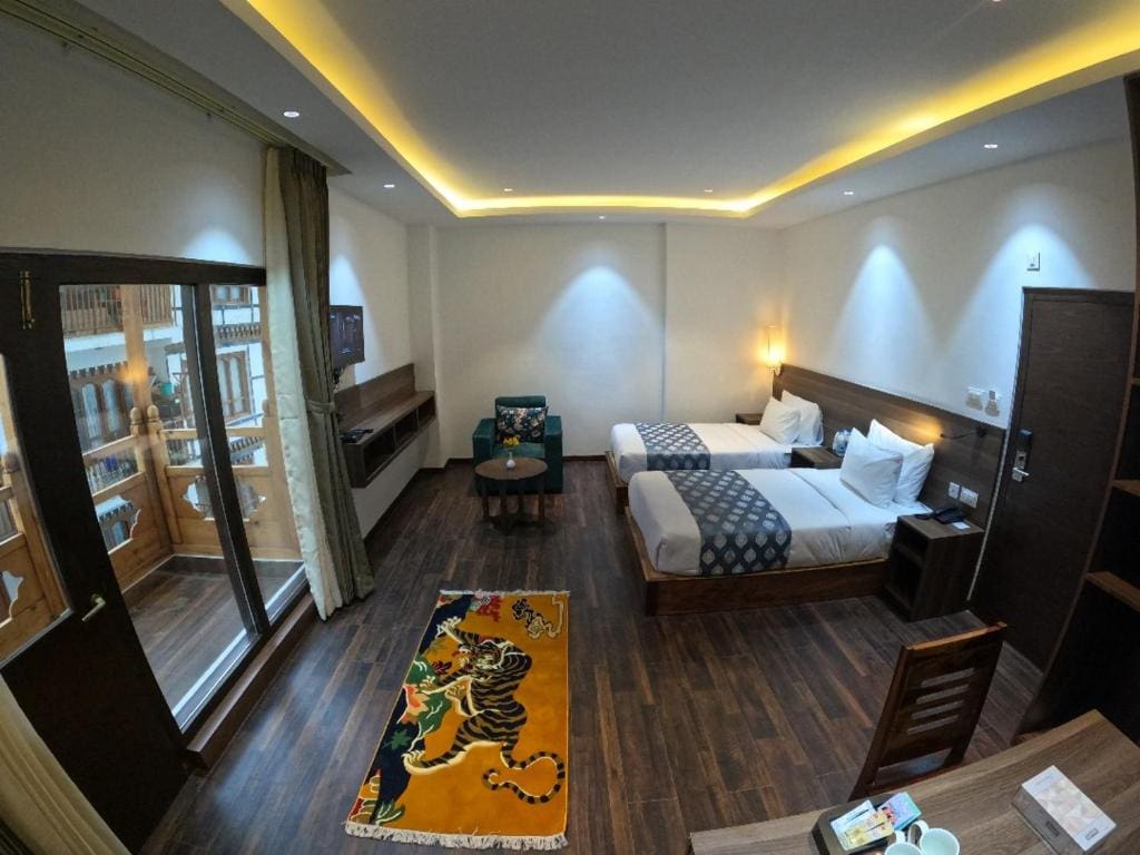 A hotel room with two single beds, a tiger print rug, a chair, a small table, a desk, and a glass door leading to a balcony; wood floors and modern lighting create a cozy atmosphere.