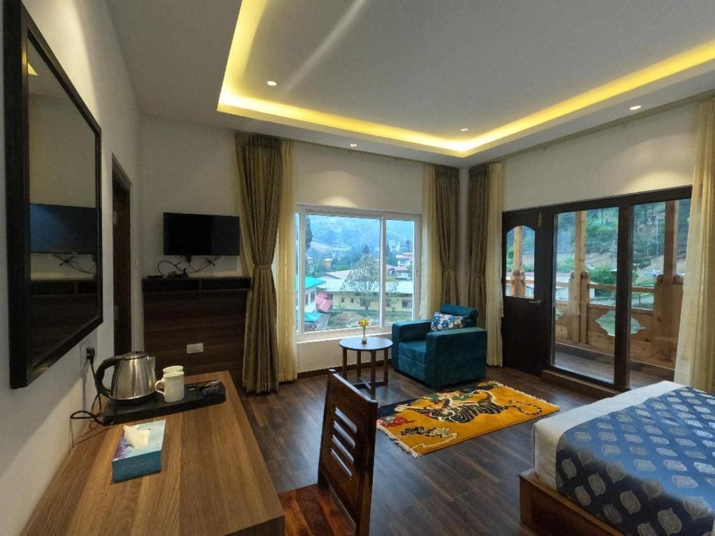A cozy hotel room features a bed, desk, TV, kettle, and a seating area by large windows with scenic mountain views. Glass doors lead to a balcony. Warm lighting and wooden flooring create an inviting atmosphere.