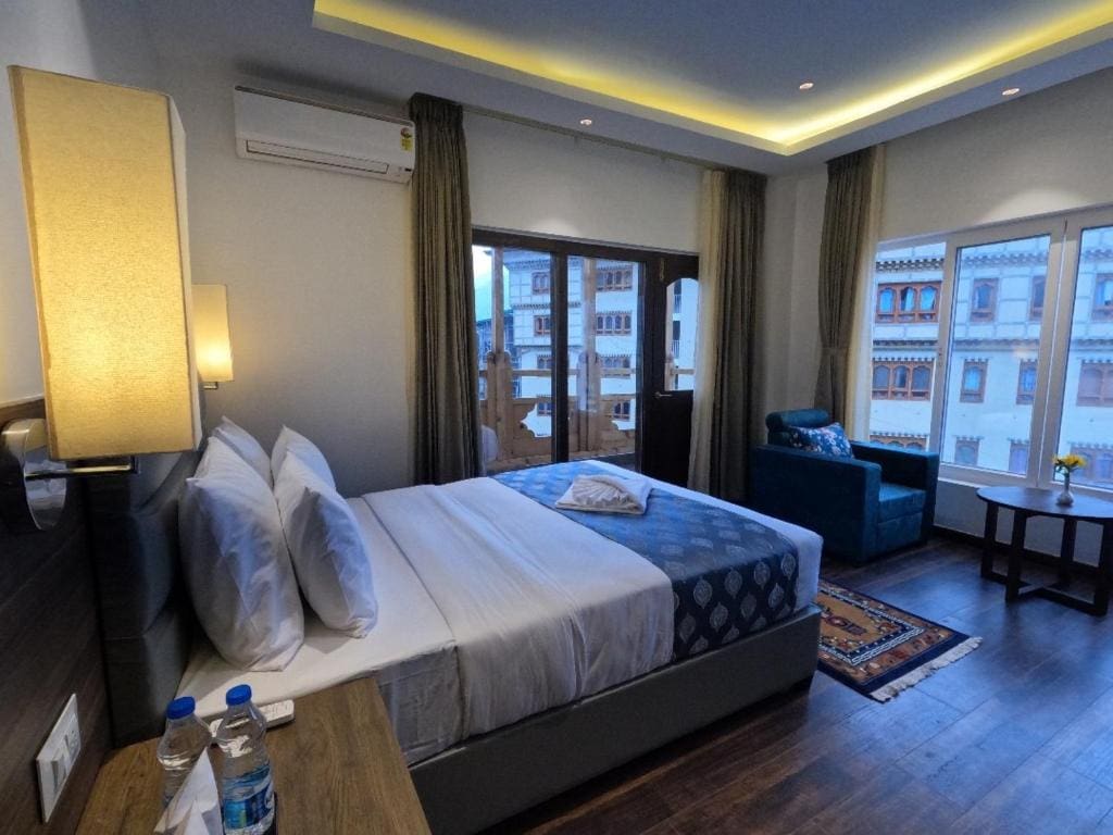 A modern hotel room with a double bed, blue armchair, side table with flowers, and large windows showing traditional-style buildings outside. Bottled water and glasses are on the wooden bedside table.
