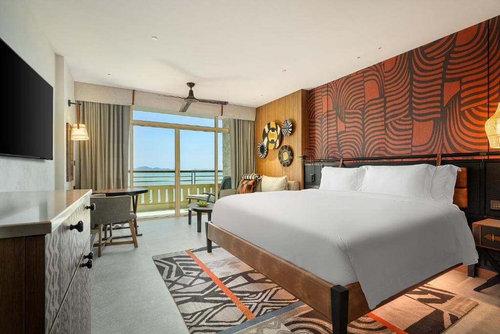 Modern hotel room with a large bed, colorful patterned accent wall, desk and chairs, and a balcony with a view of the ocean through glass doors.