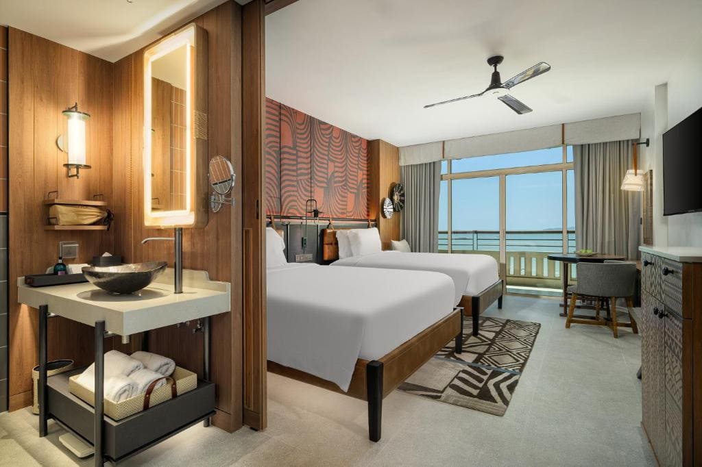 Modern hotel room with two double beds, a wall-mounted TV, a vanity sink area, a ceiling fan, a desk by the window, and a balcony with an ocean view. The decor features warm wood and patterned accents.