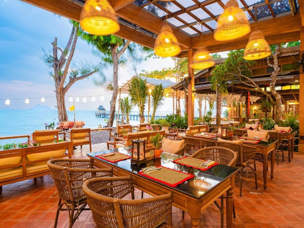 Outdoor restaurant with wicker chairs and wooden tables set for dining, under warm hanging lights. Surrounded by greenery and overlooking a calm beach with string lights and ocean in the background.